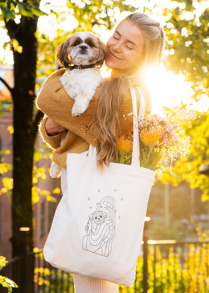 Mina står i profil og holder hunden sin. På skuldren henger ett handlenett med ett illustert motiv av Mina og hunden.