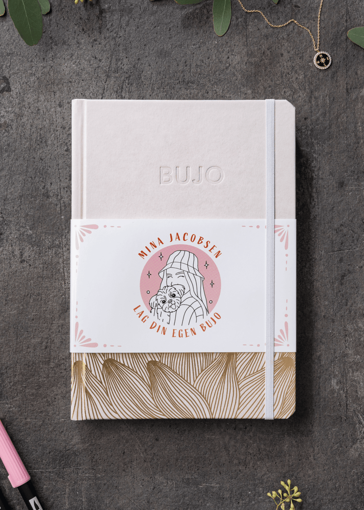 Fugleperspektiv på Minas tomme bullet journal. Boken er i lys rosa med illustrasjoner av blader på bunn i gull farge. 