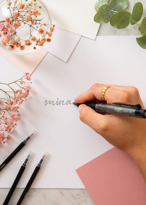 
            
                Last inn bildet i Gallery viewer, En hånd skriver ordet Mina med pennen Pilot frixon på et papir. Bordet er dekorert med papir, blomster og med påfyll til pennen. 
            
        