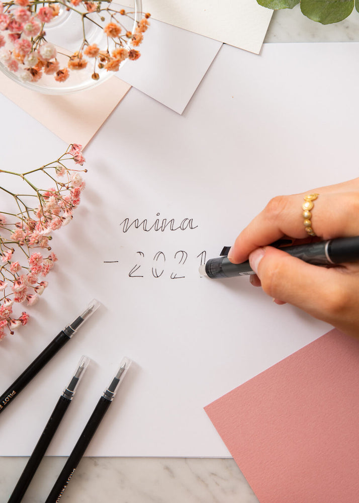 En hånd visker ut ordet Mina med pennen Pilot frixon på et papir. Bordet er dekorert med papir, blomster og med påfyll til pennen. 