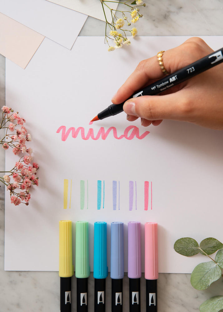 
            
                Last inn bildet i Gallery viewer, En hånd skriver ordet Mina med en rosa penseltusj på et papir. Bordet er dekorert med papir og blomster.
            
        