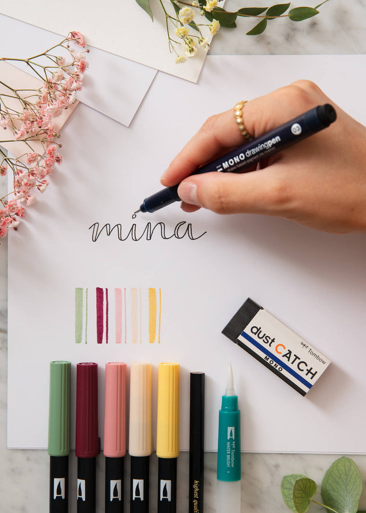 
            
                Last inn bildet i Gallery viewer, En hånd skriver ordet Mina med en svart penn på et papir. Fargepenner, viskelær og blyant ligger ved siden av. 
            
        